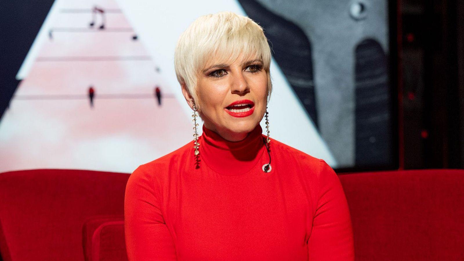 Pasión Vega, cantante, con cabello rubio platino y jersey rojo, sentada frente a un fondo con pentagrama y figuras.