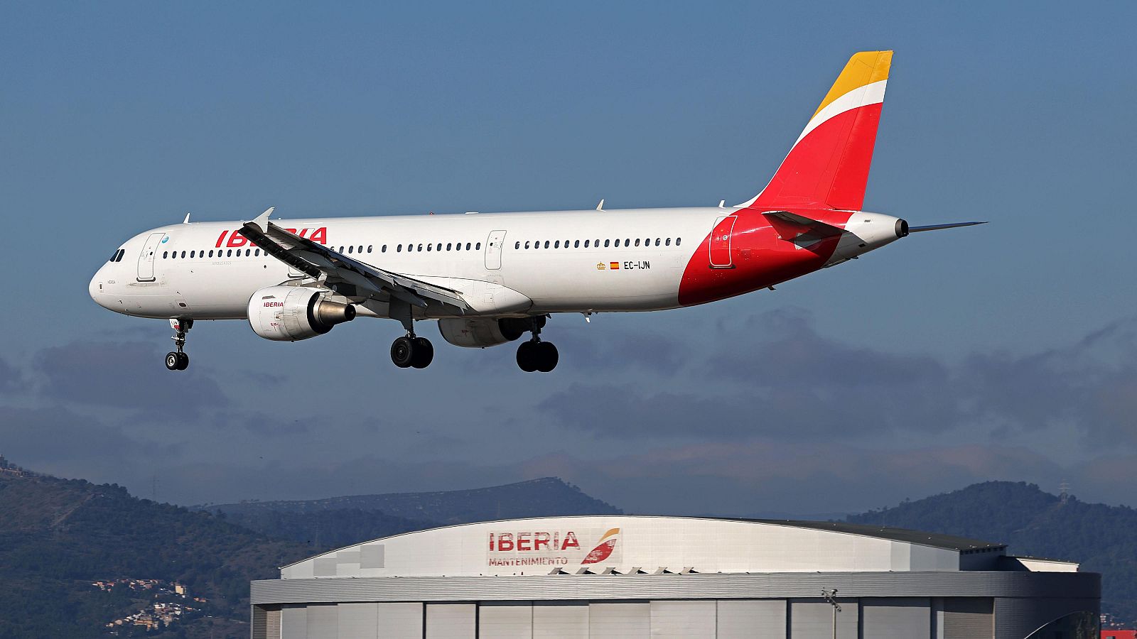 Avión de Iberia EC-LJN en vuelo con tren de aterrizaje desplegado, sobre un edificio de Iberia Mantenimiento.