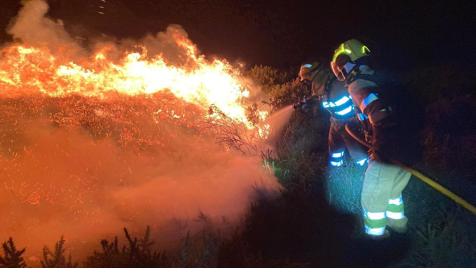 Bomberos combaten un incendio nocturno con agua y mangueras. El fuego ilumina la escena con llamas naranjas y amarillas, mientras el humo se eleva. Uno de los bomberos lleva un casco amarillo.
