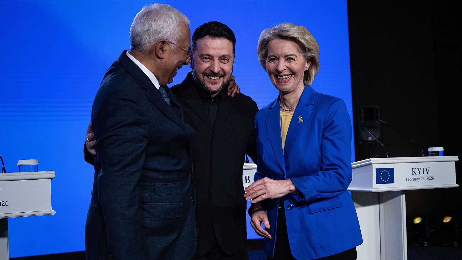 Volodymyr Zelenskyy, Antonio Costa y Ursula von der Leyen sonríen en un evento. Zelenskyy abraza a Costa, mientras von der Leyen luce un blazer azul y un pin amarillo. Se ven podios con el logo de la UE y "KYIV".