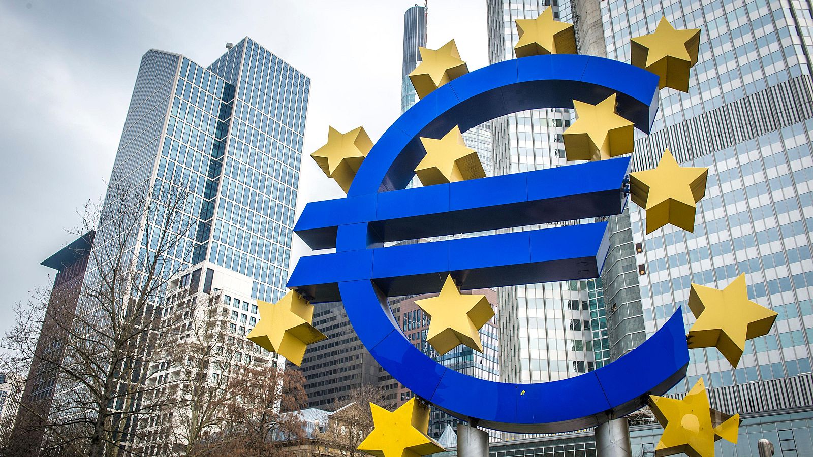 Escultura tridimensional del símbolo del euro en azul intenso con estrellas doradas, rodeada de rascacielos y cielo nublado.