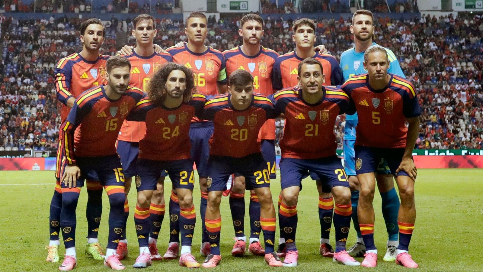 Jugadores de la selección española posan en el césped con su uniforme rojo, azul y amarillo, luciendo escudos y logos de Adidas. Se distinguen números y detalles en sus camisetas y botas.