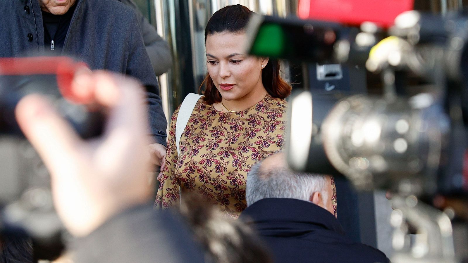 Primer plano de Elisa Mouliaá, con expresión seria, rodeada de cámaras y micrófonos tras una comparecencia. Lleva blusa dorada y fina cadena.