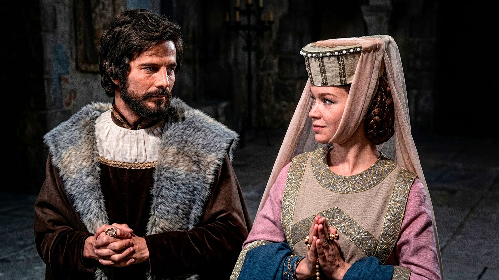 José María Prada y Victoria Vera como Rey y Reina en "El juglar y la reina" (RTVE), con vestimenta de época y expresión solemne en un interior de piedra.