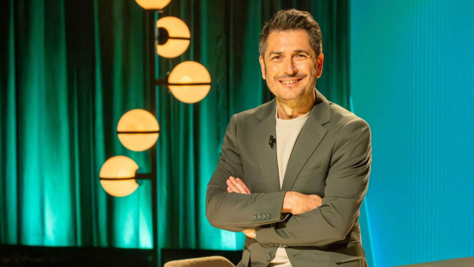 Carlos del Amor, periodista de RTVE, posa sonriente con chaqueta verde y camiseta clara, brazos cruzados, ante un fondo de cortinas verdes y pared azul turquesa con lámpara decorativa.
