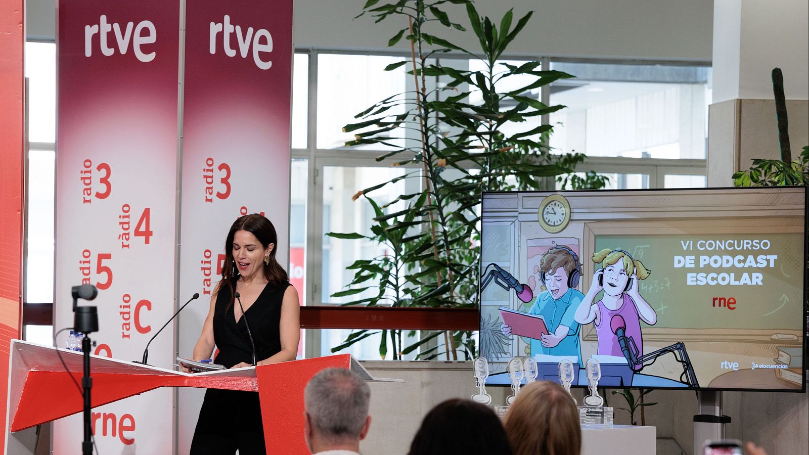 Paula Aller habla en un podio con el logo "rne". Detrás, banners de "rtve" y emisoras de RNE. Una pantalla muestra el "VI CONCURSO DE PODCAST ESCOLAR" con niños y logos.