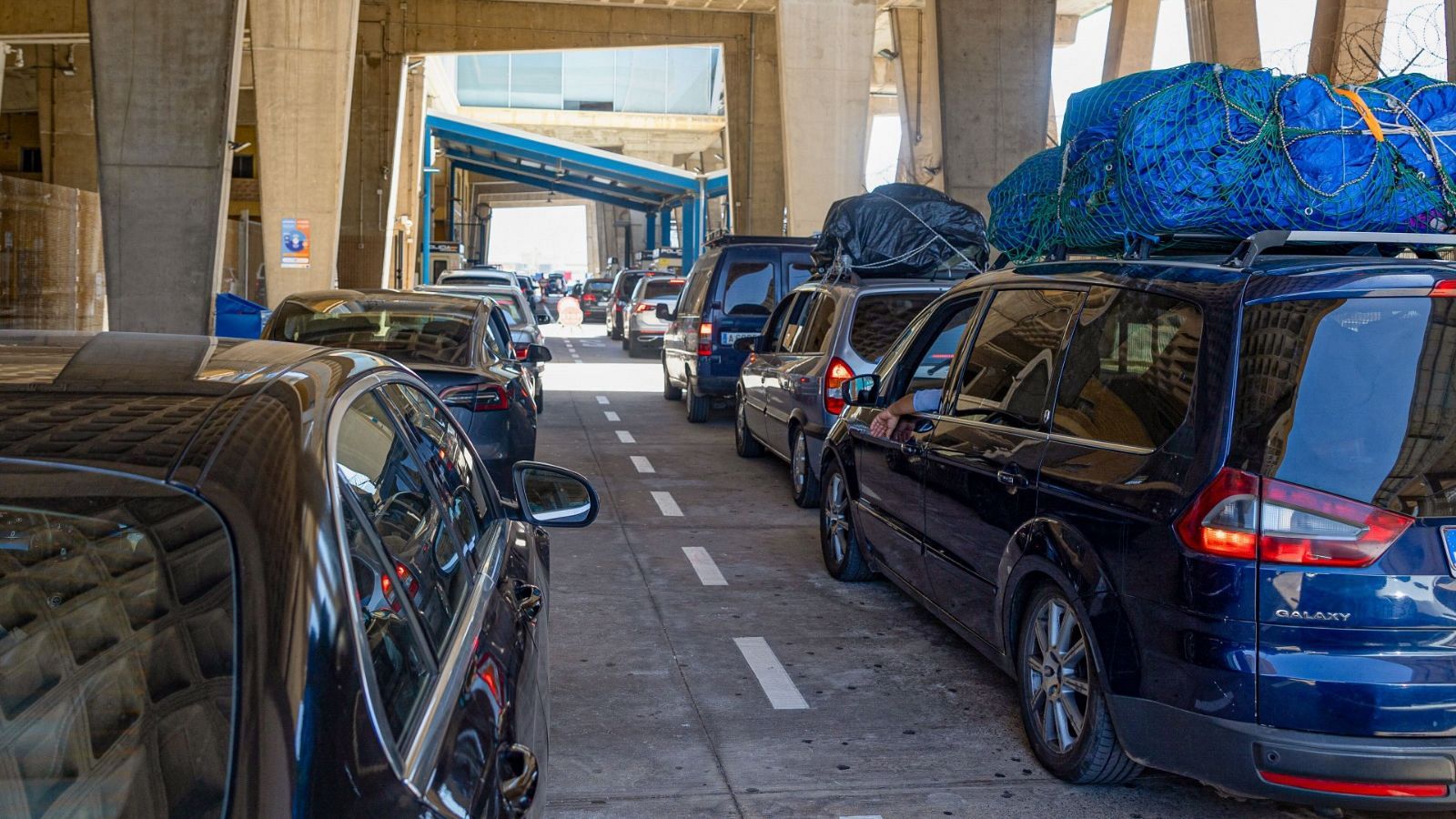 Numerosos vehículos, muchos con equipaje en sus techos, avanzan lentamente bajo una estructura de hormigón. Se distingue un Ford Galaxy en la fila.