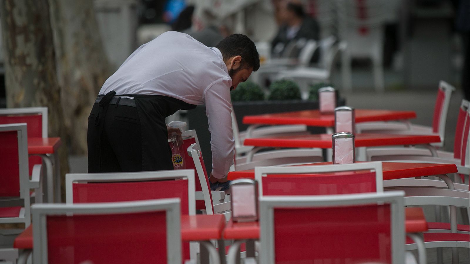 Camarero con uniforme y delantal negro limpia silla en terraza con mobiliario rojo y blanco. Sostiene botella de spray. Día.