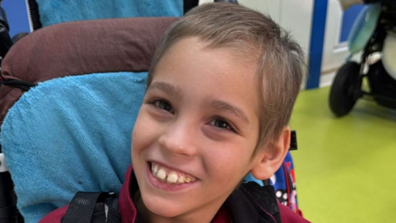 Adrián, un niño de Polán, Castilla-La Mancha, sonríe a la cámara mientras está sentado en una silla especializada. Viste una prenda granate y su pelo es corto y despeinado.