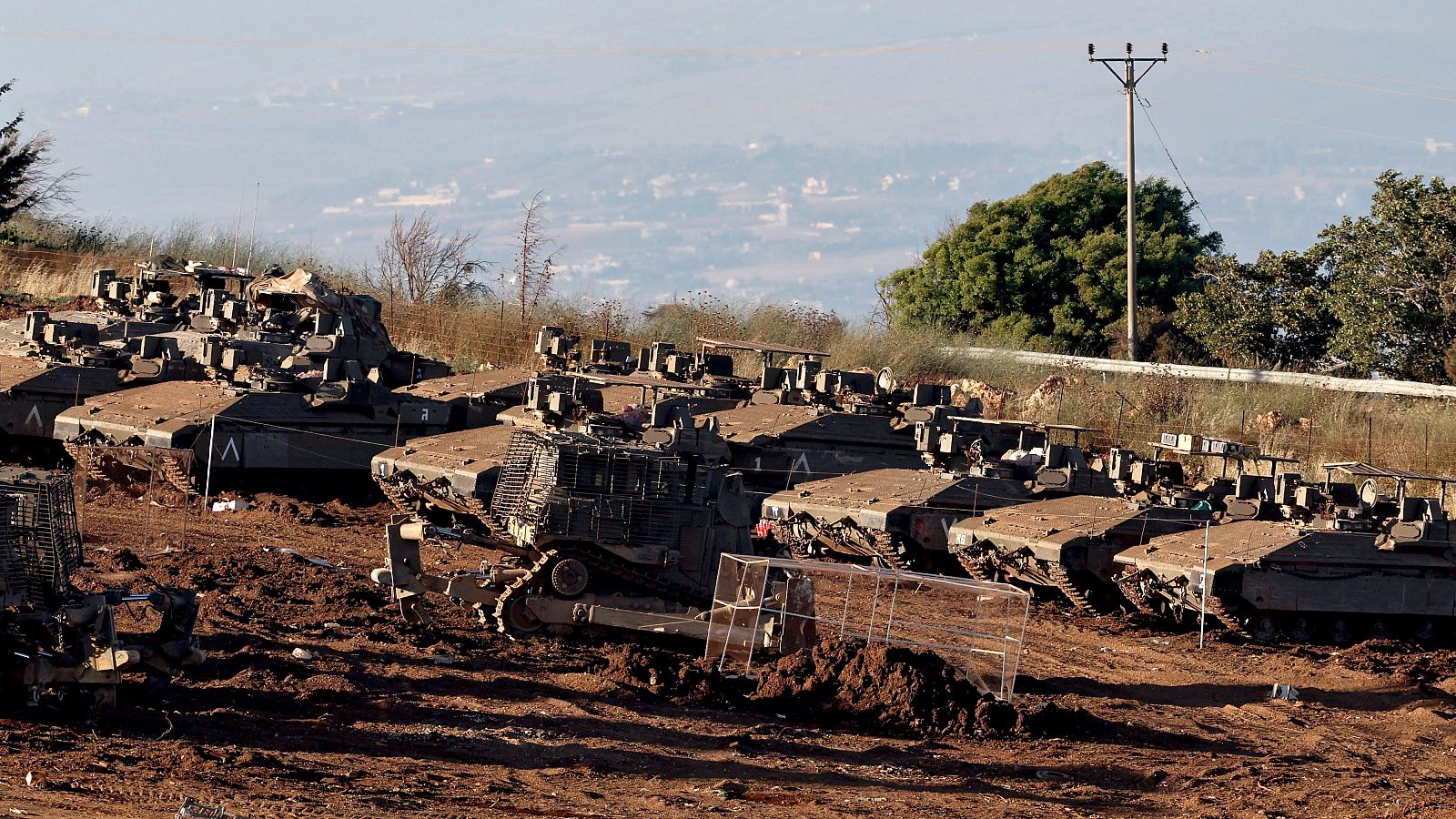 Numerosos tanques israelíes se encuentran estacionados en un terreno irregular en la frontera con Líbano. Se aprecian marcas blancas en 'V' en algunos vehículos. El fondo muestra colinas y una zona urbana.