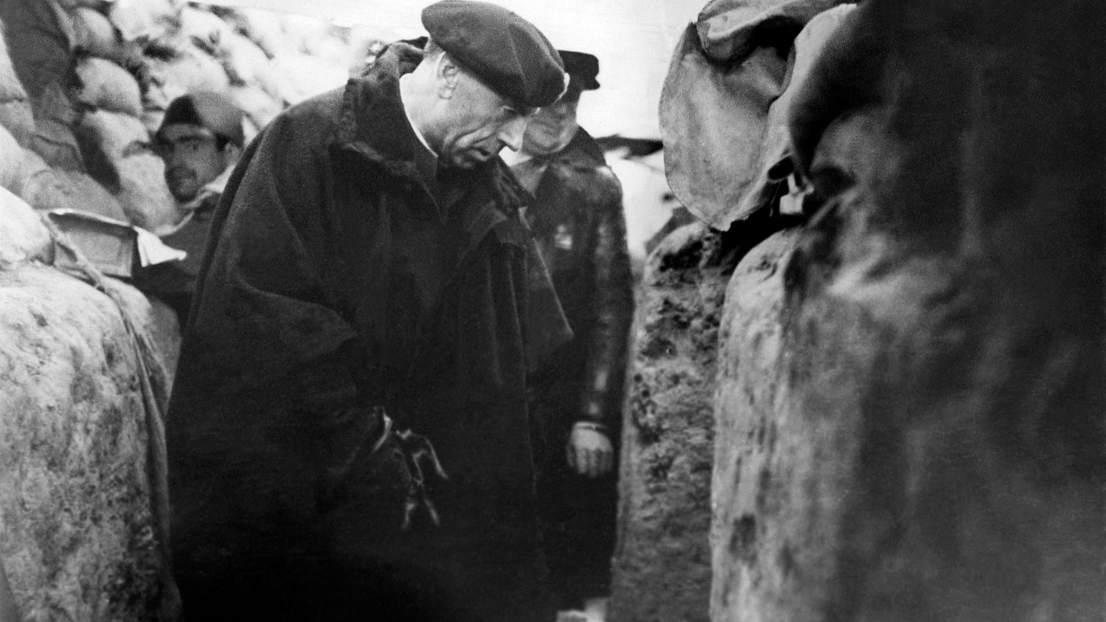 Antonio Ortega en las trincheras de Madrid en 1936, con expresión reflexiva, rodeado por sacos de arena y acompañado por otros hombres.