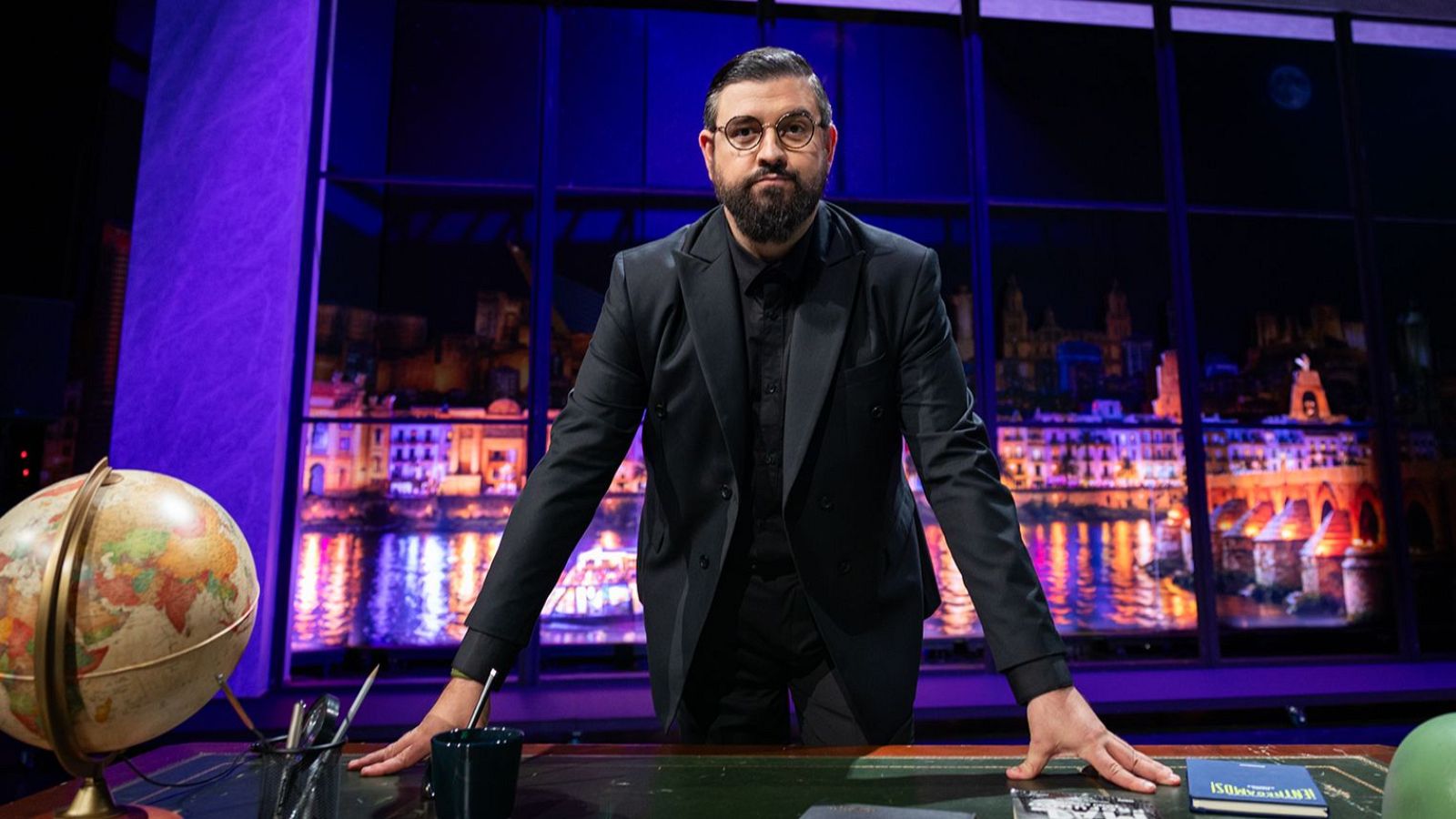 Manu Sánchez, con traje oscuro y gafas, posa en un estudio con un globo terráqueo y un libro sobre el escritorio. Al fondo, una ciudad nocturna iluminada a través de una ventana.