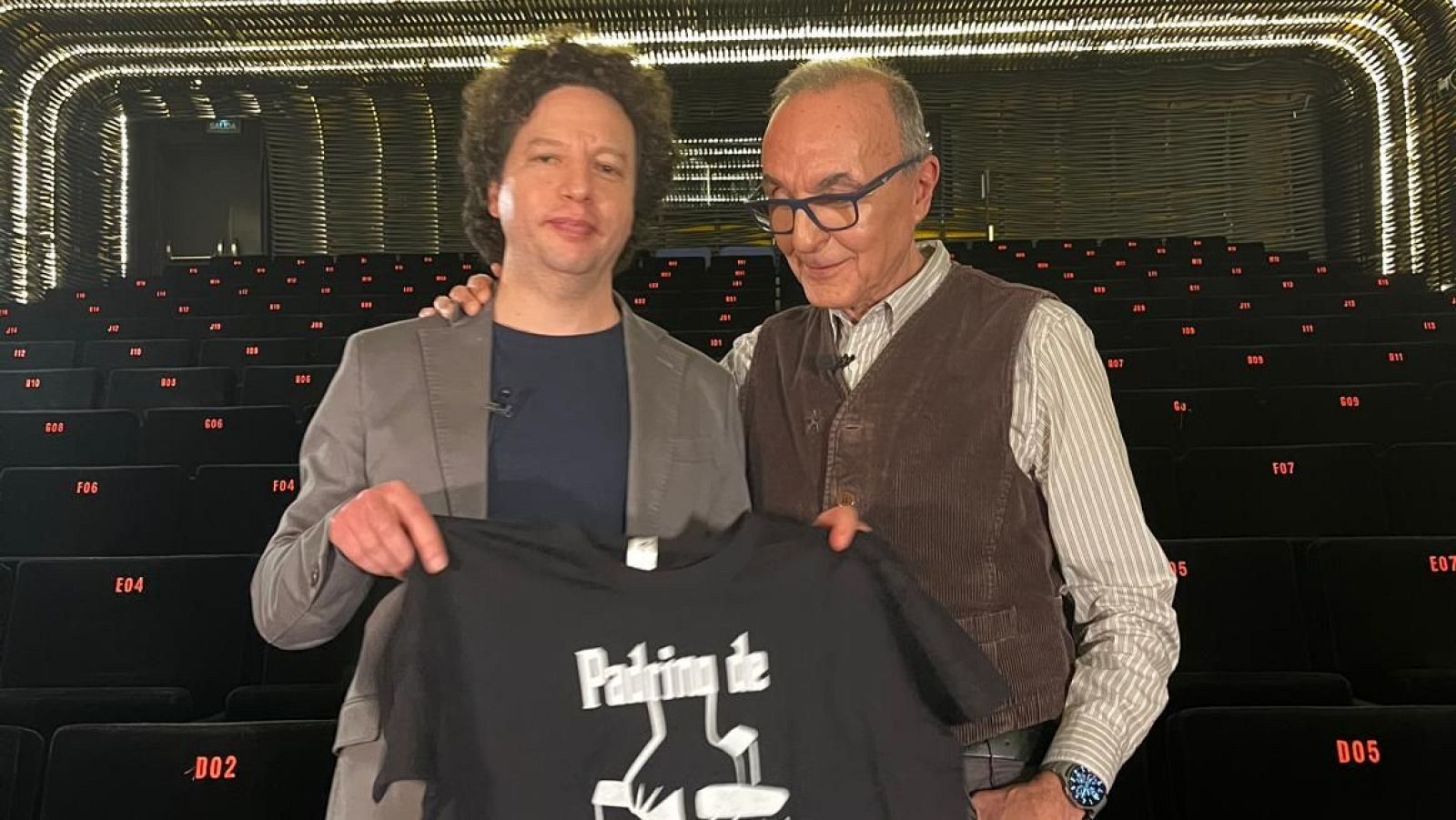 Michel Franco y Gerardo Sánchez, ambos con micrófonos, posan en una sala de cine vacía sosteniendo una camiseta negra con el texto "Padrino de" y un diseño cinematográfico.