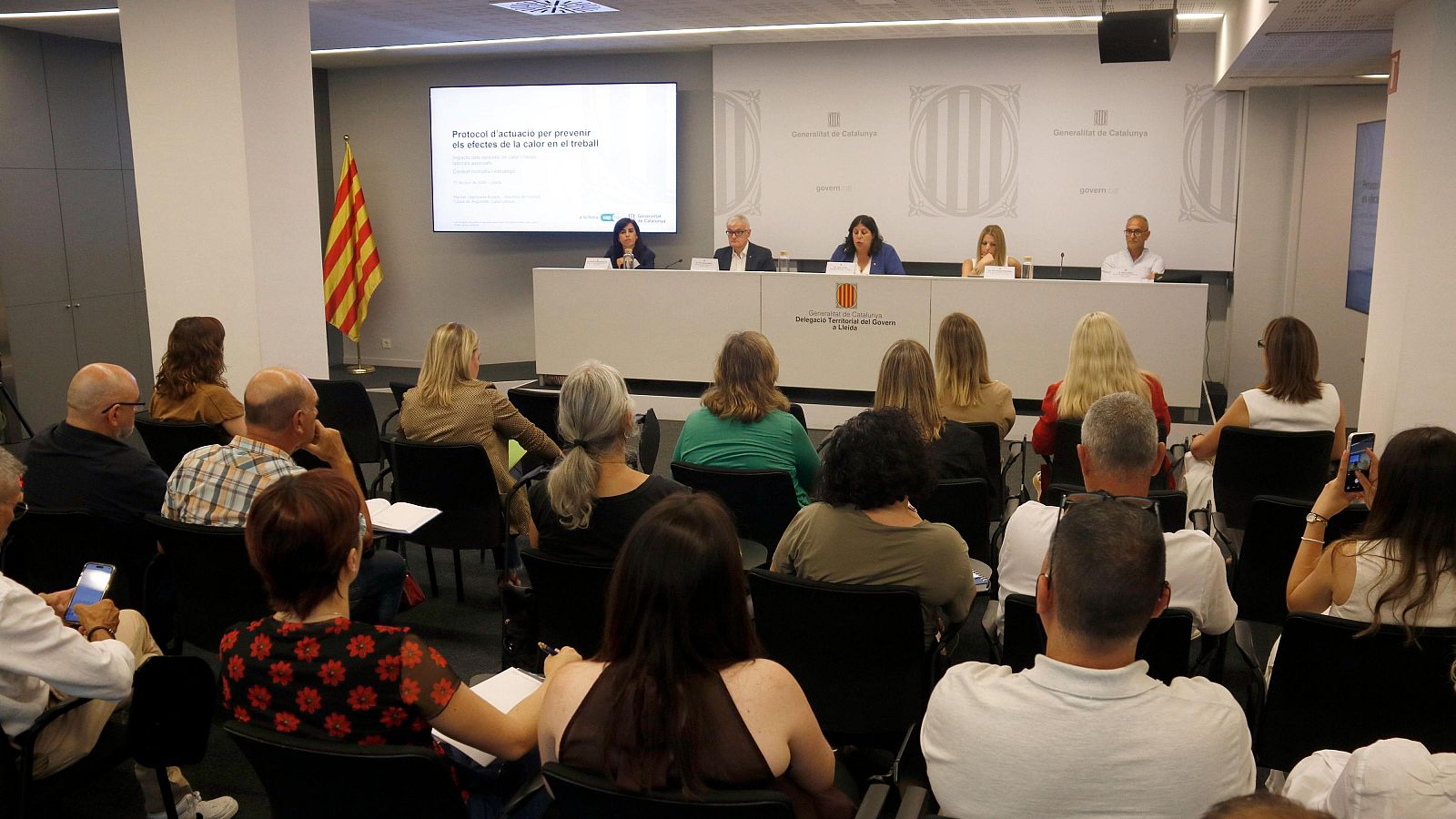 Evento institucional sobre el calor en el trabajo en Lleida, con ponentes y audiencia en una sala de conferencias. Se visualiza un protocolo de actuación en una pantalla.
