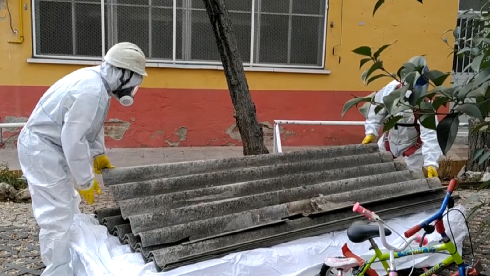 Treballadors amb equips de protecció manipulen plaques de fibrociment ondulades. Al fons, un edifici groc i vermell. A primer pla, una bicicleta infantil i una porteria de futbol.
