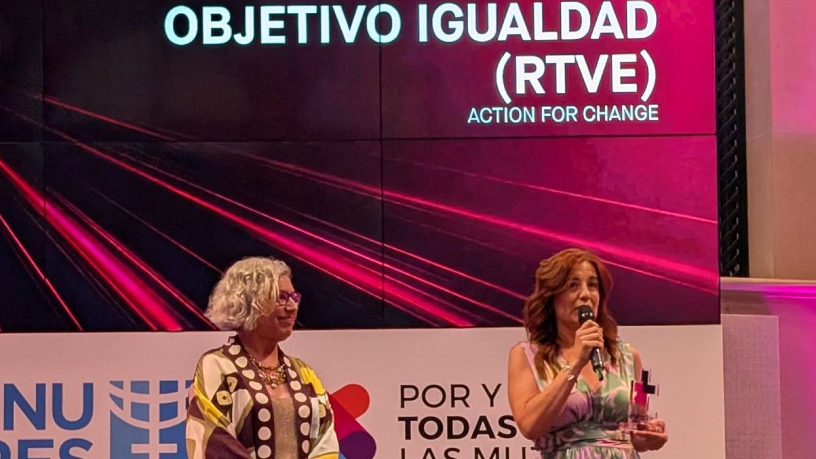 Carolina Pecharromán y Paola Guerra de la Fuente reciben un premio en el escenario de "Objetivo Igualdad" de RTVE, con una pantalla de fondo y un trofeo.