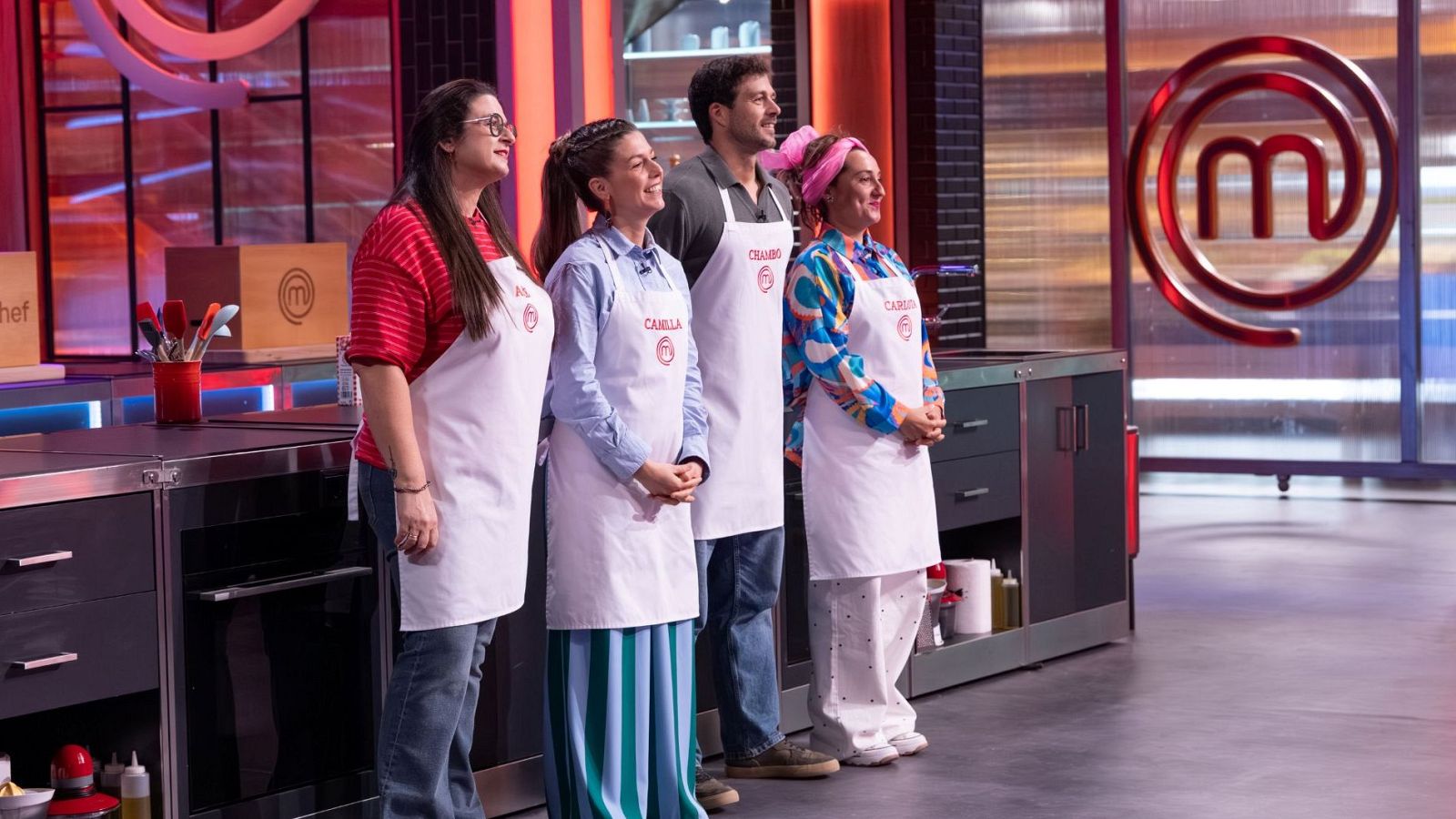 Annie, Camilla, Chambo y Carlota, finalistas de MasterChef, posan con sus delantales blancos en un estudio de cocina. El logo de MasterChef se ilumina al fondo.