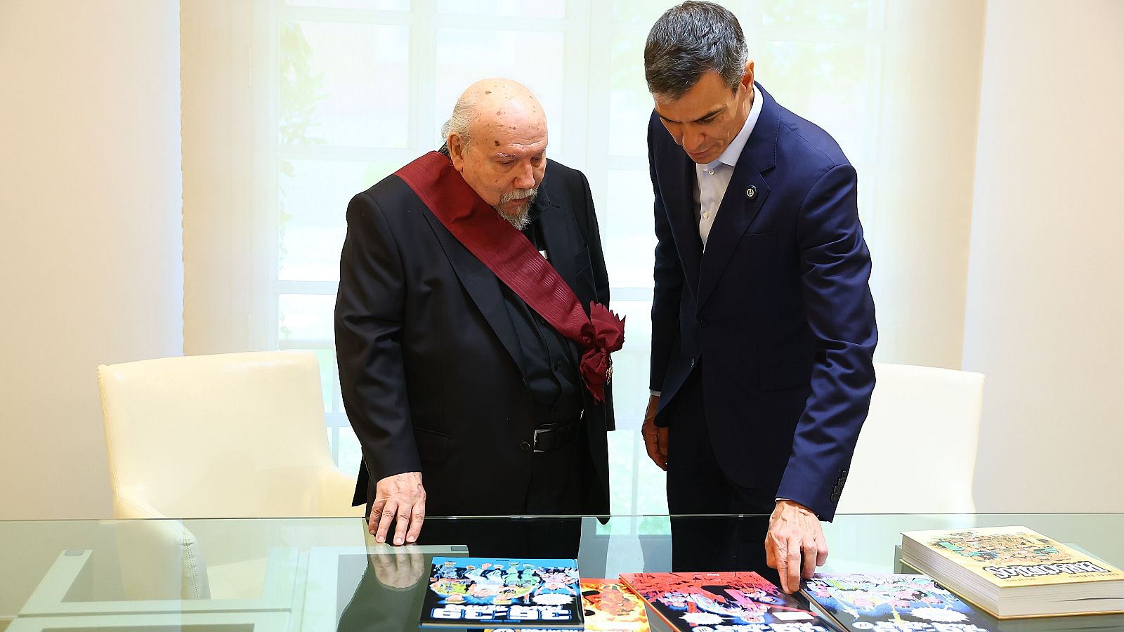 Pedro Sánchez entrega la Gran Cruz de Alfonso X el Sabio a Carlos Giménez mientras ambos observan cómics sobre una mesa de cristal.