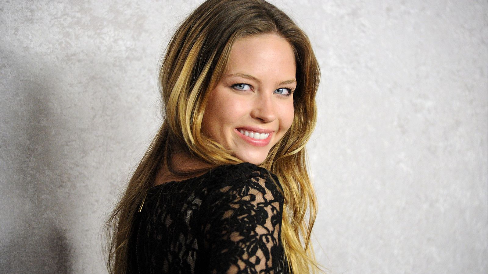 Retrato de Daveigh Chase con cabello largo y ondulado, ojos azules y una amplia sonrisa, vestida con un top de encaje negro. Fondo gris claro.