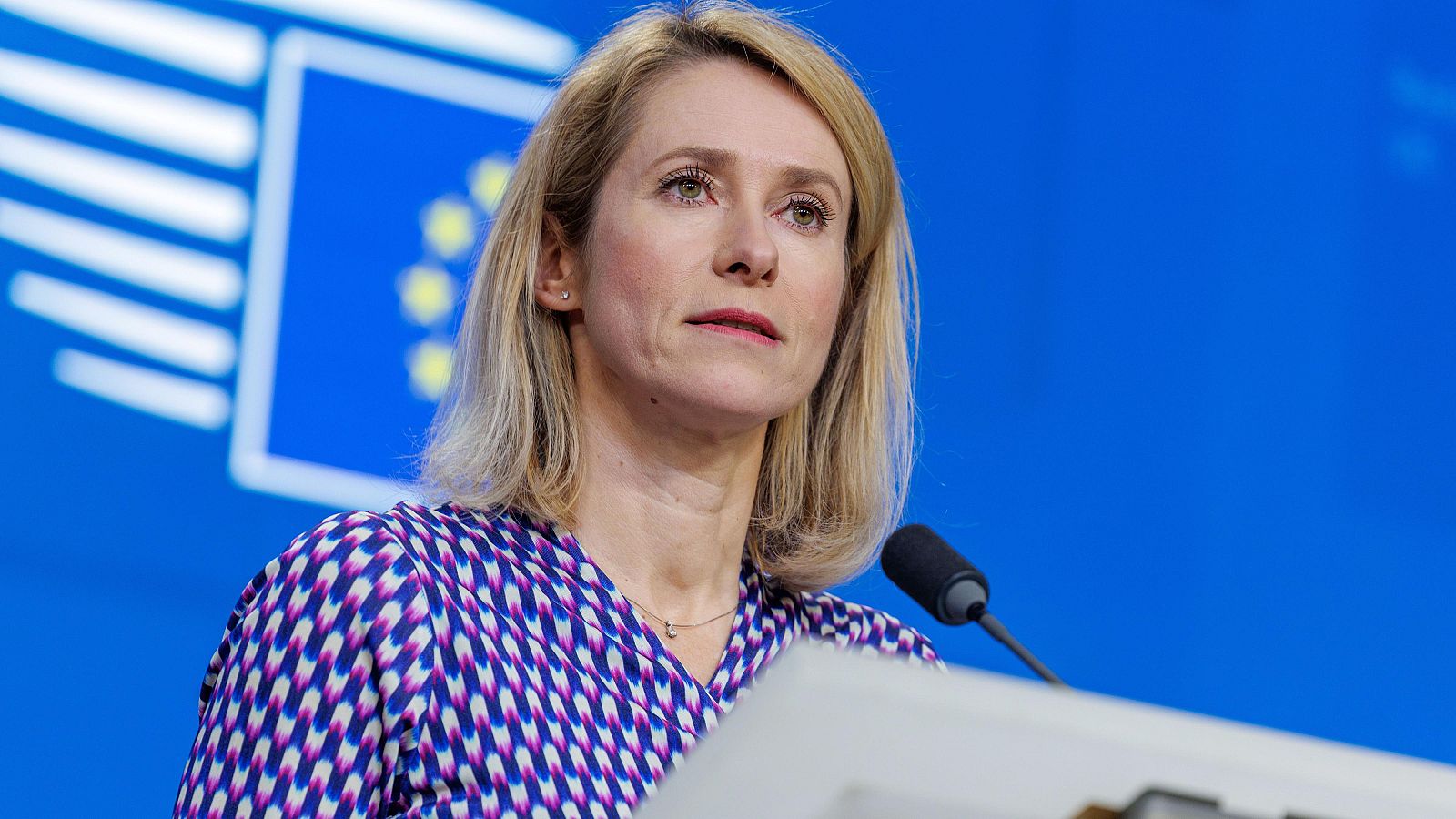 En la imagen, Kaja Kallas, con cabello rubio y una blusa estampada, habla ante un micrófono. Se aprecia la bandera de la Unión Europea al fondo, sugiriendo un evento oficial.