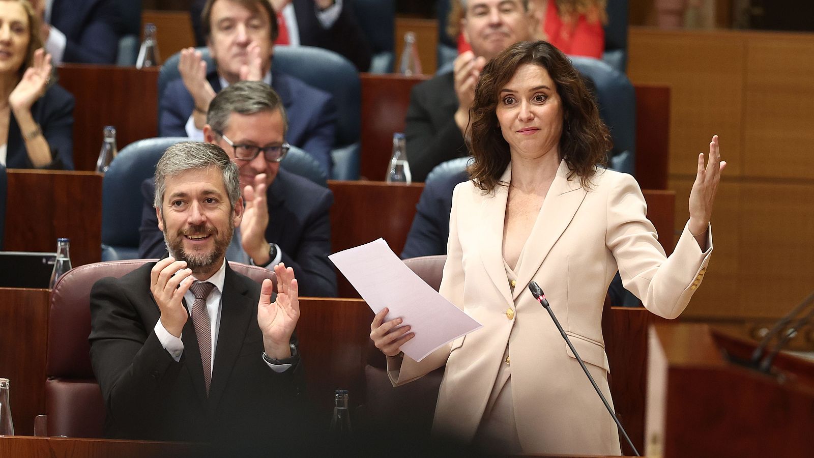 Isabel Díaz Ayuso, presidenta de la Comunidad de Madrid, en una sesión plenaria en la Asamblea de Madrid, de pie y hablando. A su izquierda, un hombre aplaude.