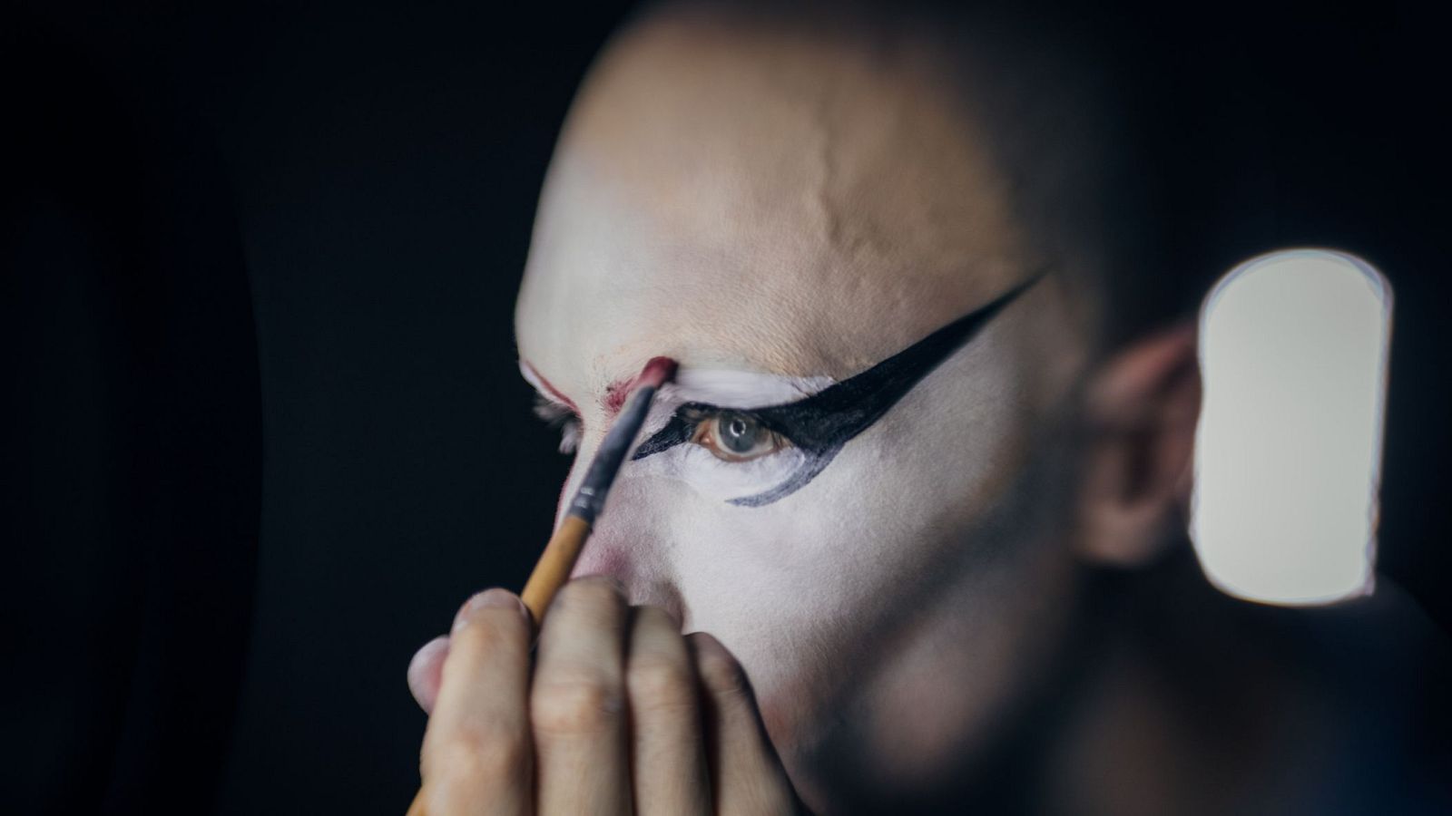 Rostro calvo con maquillaje artístico: base pálida, delineado negro dramático y acento rojo en el párpado. Una mano aplica el color con un pincel.