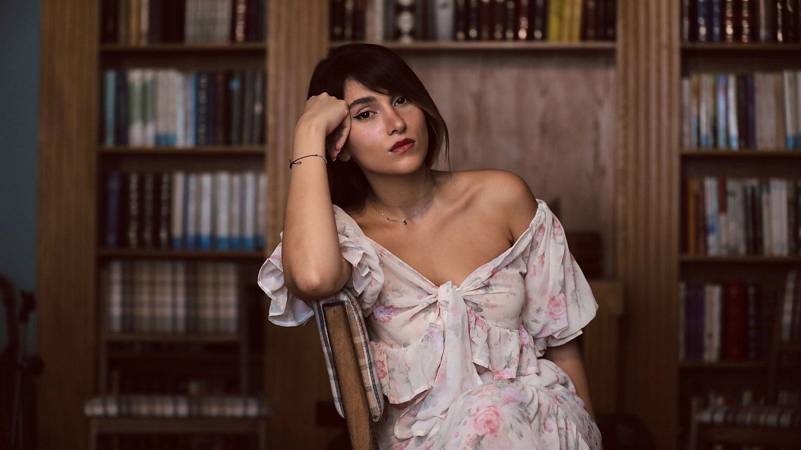Retrato de la artista iraní Parastoo Ahmadi, con vestido floral y pose reflexiva, en un entorno de estanterías llenas de libros.