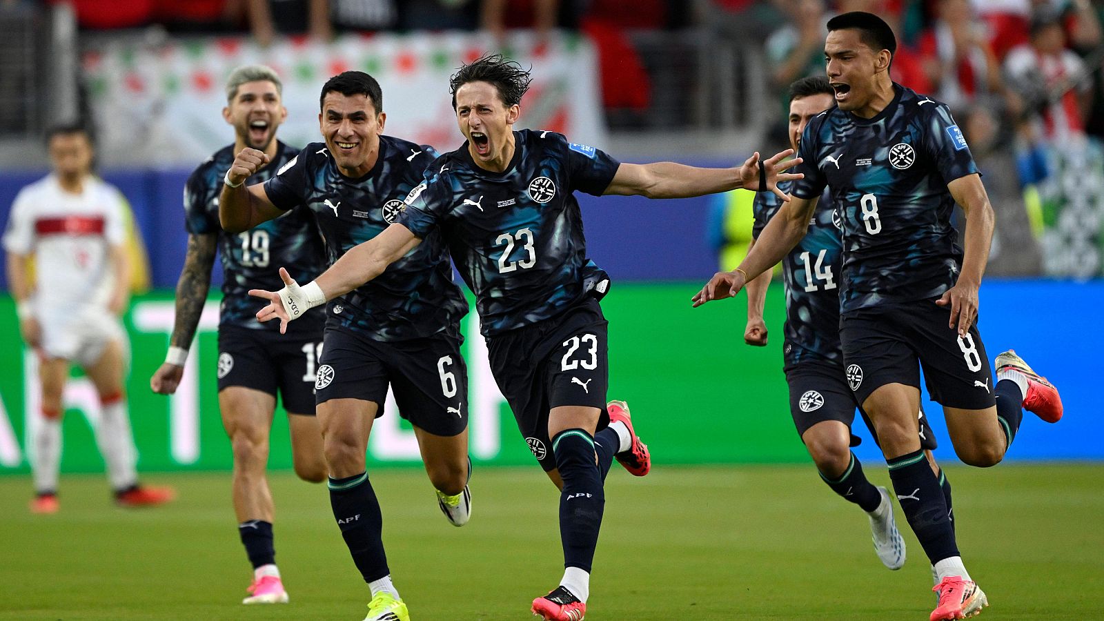 Futbolistas paraguayos celebran un gol en un partido. El jugador 23 es el centro de la euforia, con compañeros celebrando a su alrededor en el campo de césped.
