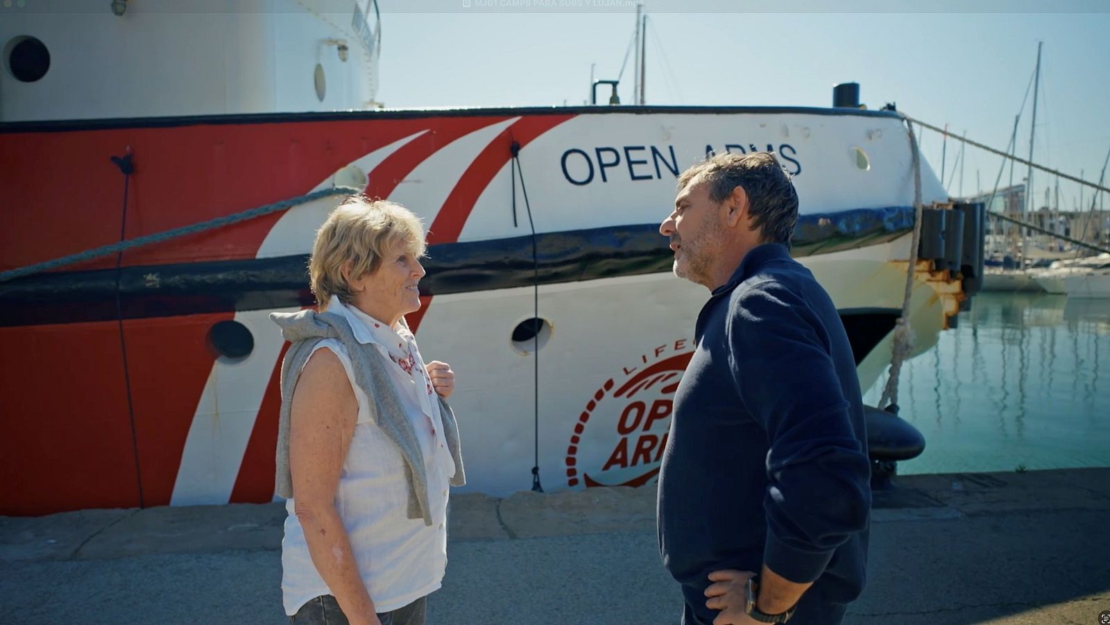 Mercedes Milá y Óscar Camps en un muelle soleado, junto al barco "OPEN ARMS".