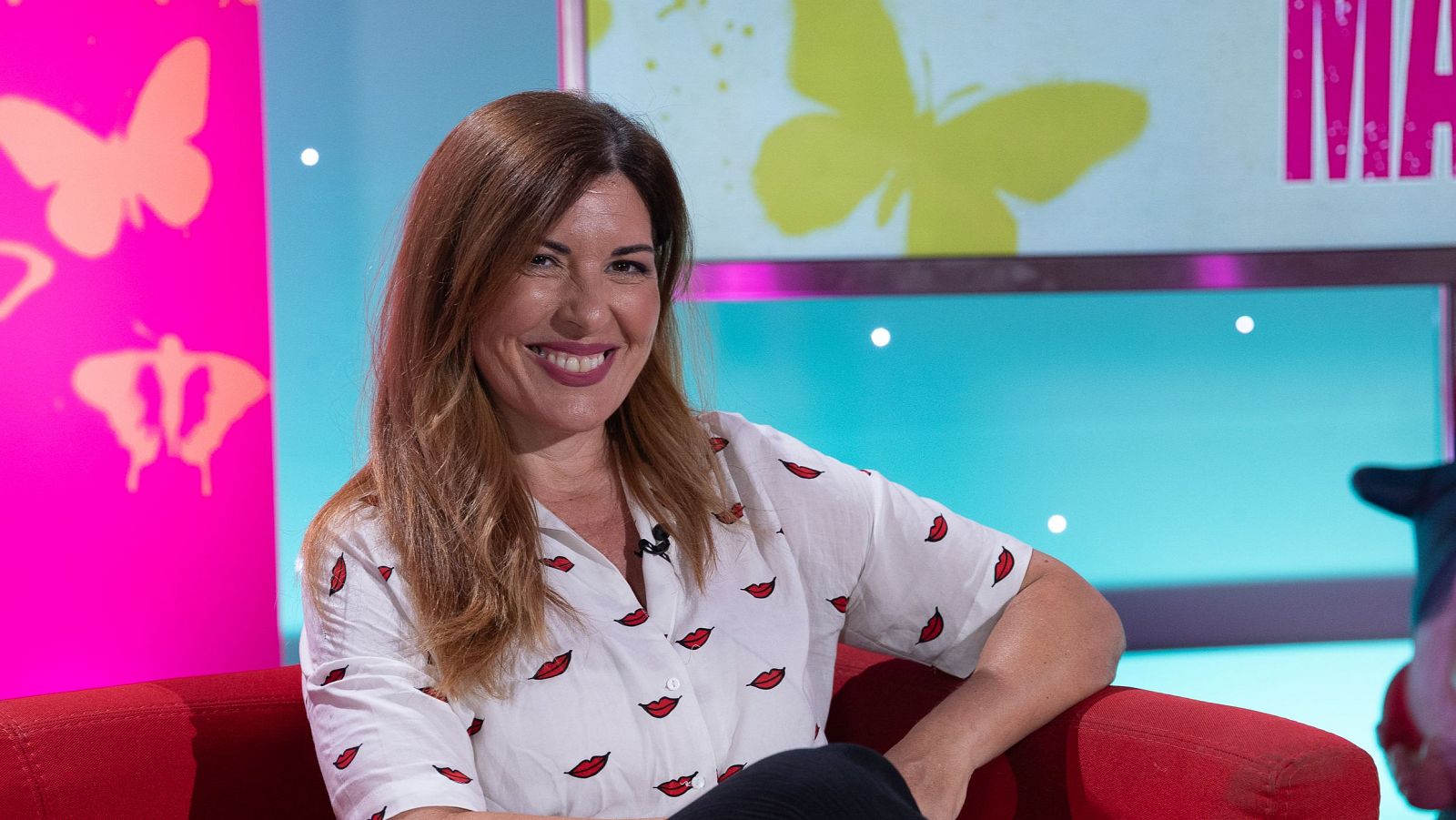 Patricia Espejo, sonriente y vestida con camisa blanca estampada, posa en un sofá rojo ante un fondo de estudio con mariposas y el texto "MA".