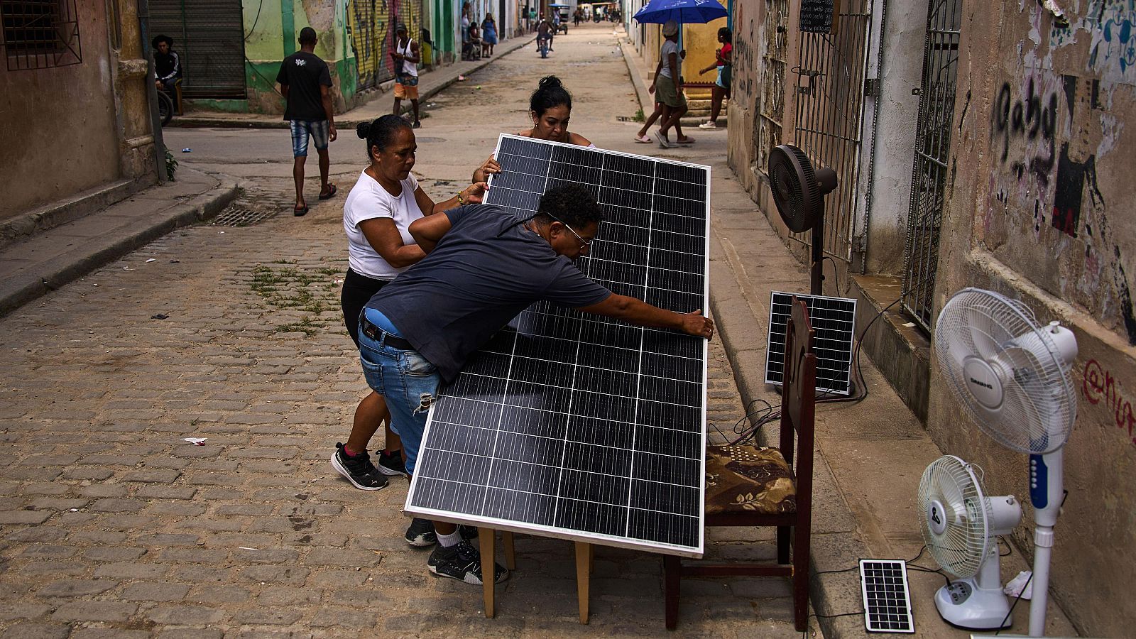 Personas instalan un panel solar en una calle adoquinada, rodeados de otros paneles, ventiladores y cables. Se aprecia un grafiti con la palabra 'gaby' en una pared.