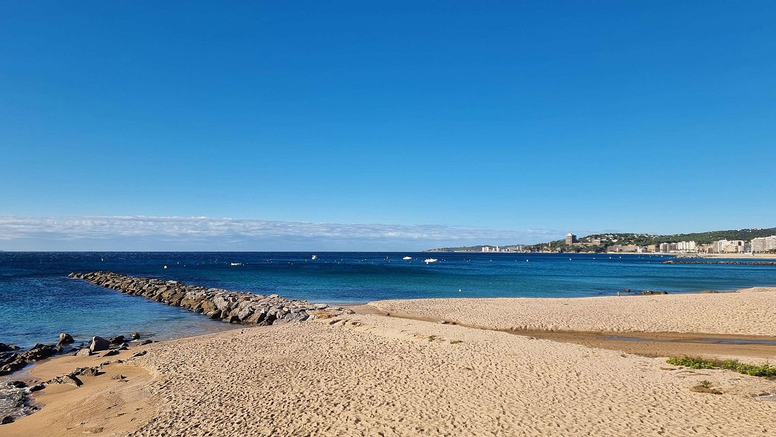 Paisaje costero con playa de arena, mar azul, riachuelo, formaciones rocosas, rompeolas y ciudad en la distancia bajo un cielo despejado.