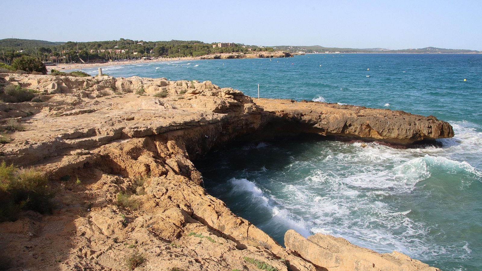 Paisaje costero con rocas, mar turquesa, playa con sombrillas y vegetación en un día soleado.