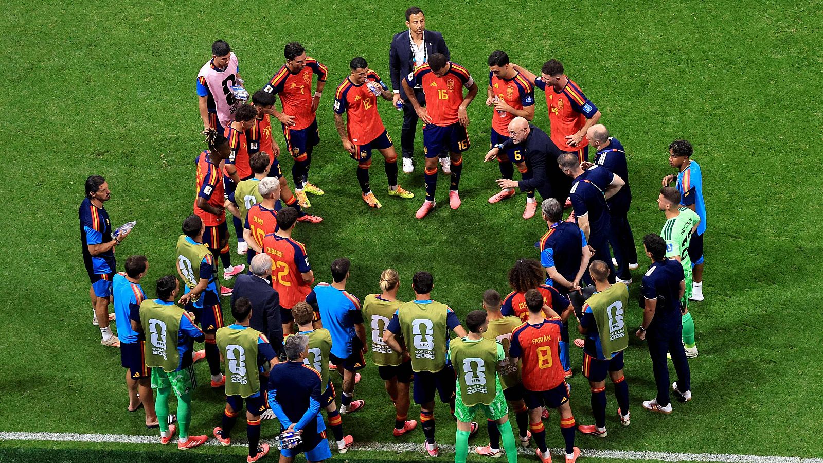 Luis de la Fuente, seleccionador español, da instrucciones a jugadores en un campo de fútbol. Se observa una pausa de hidratación con miembros del cuerpo técnico.
