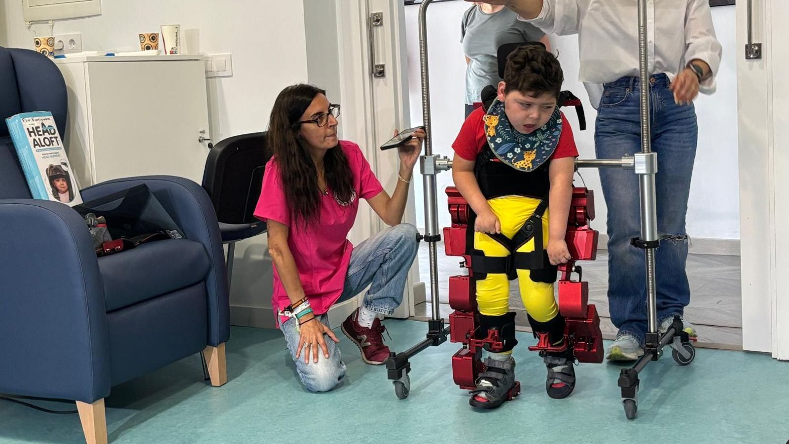 Odín, un niño con exoesqueleto robótico Atlas 2030, camina asistido por dos adultos en un centro terapéutico. Lleva camiseta roja y babero estampado. La sala es luminosa, con paredes blancas y suelo azul.