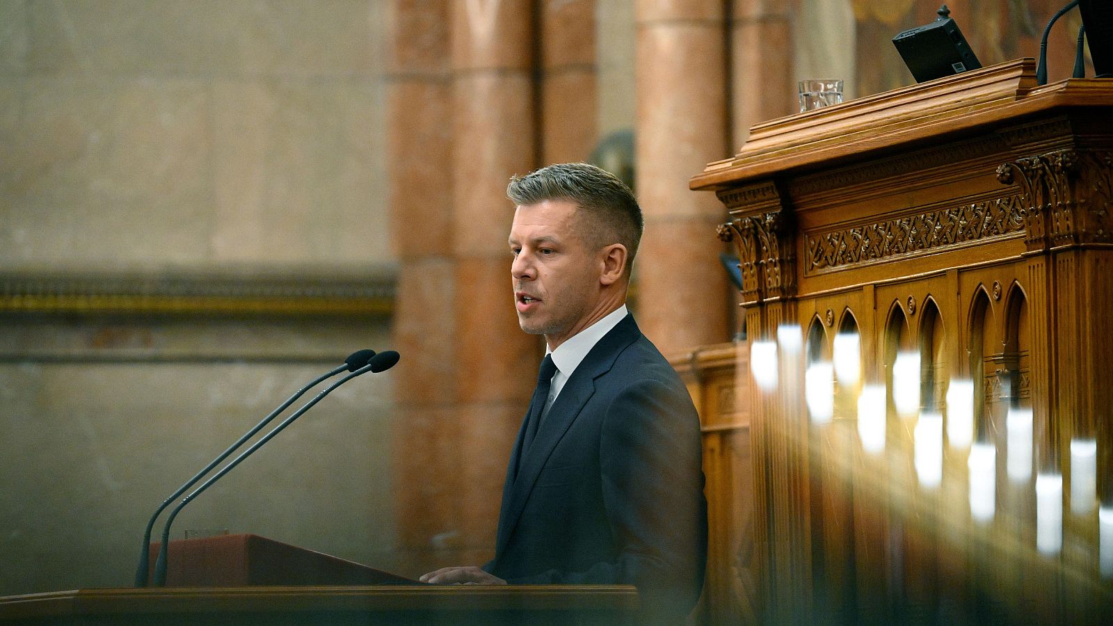 Primer plano de Peter Magyar, primer ministro húngaro, hablando en el Parlamento. Se observa un atril de madera con micrófonos y un vaso de agua.