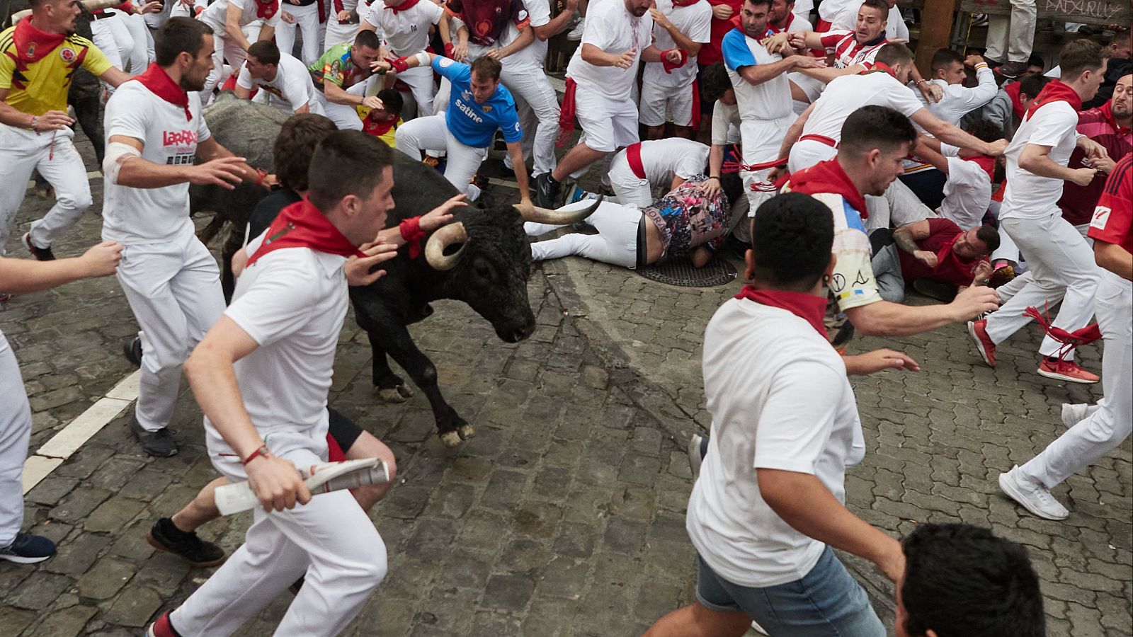 Un toro negro avanza entre corredores vestidos de blanco y rojo en un encierro de San Fermín. La escena muestra el caos y el peligro con personas corriendo y otras en el suelo.