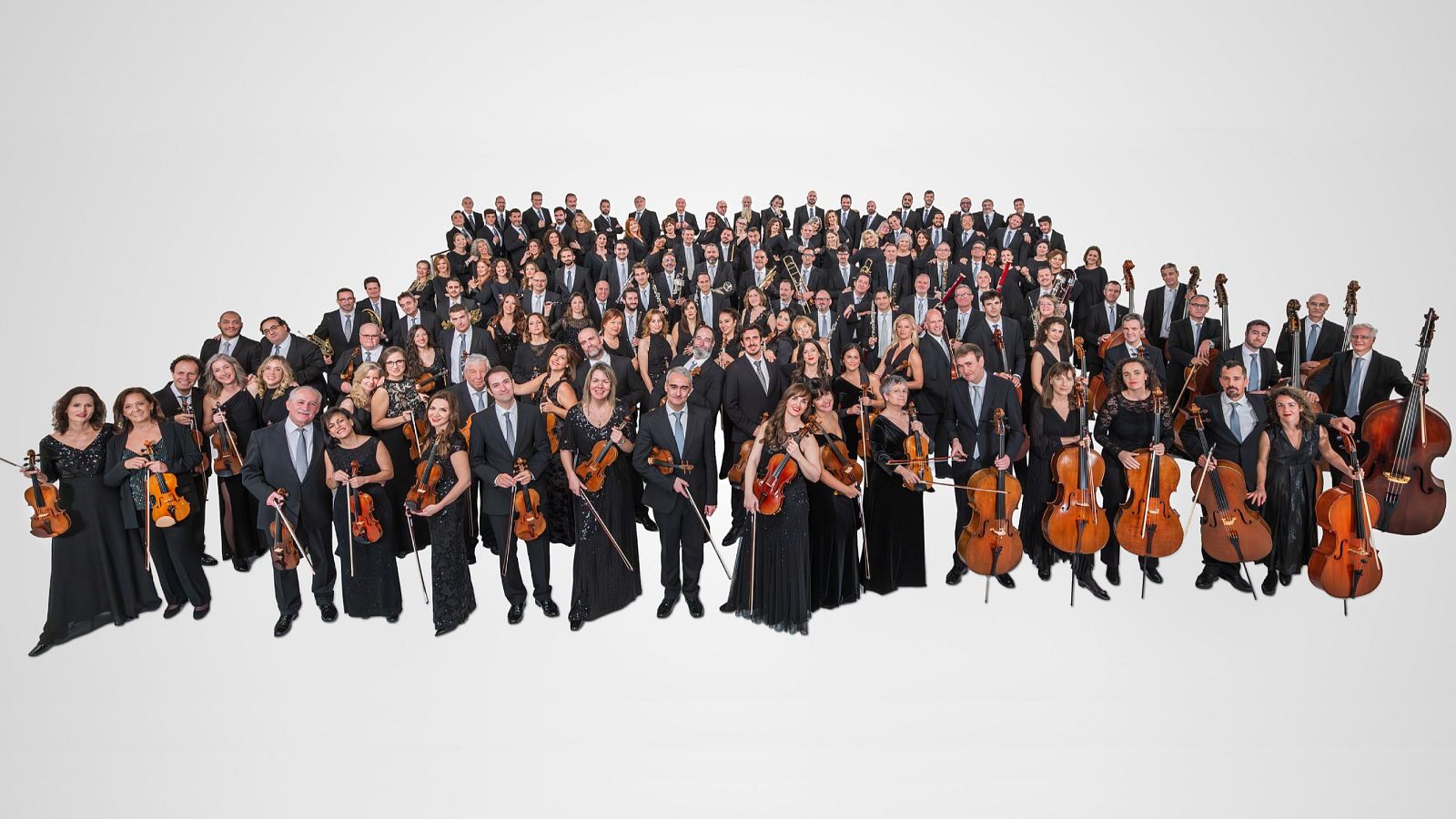 Retrato formal de la Orquesta y Coro de RTVE, con 150-200 miembros en filas escalonadas. Predominan instrumentos de cuerda. Hombres con trajes oscuros y mujeres con vestidos negros.