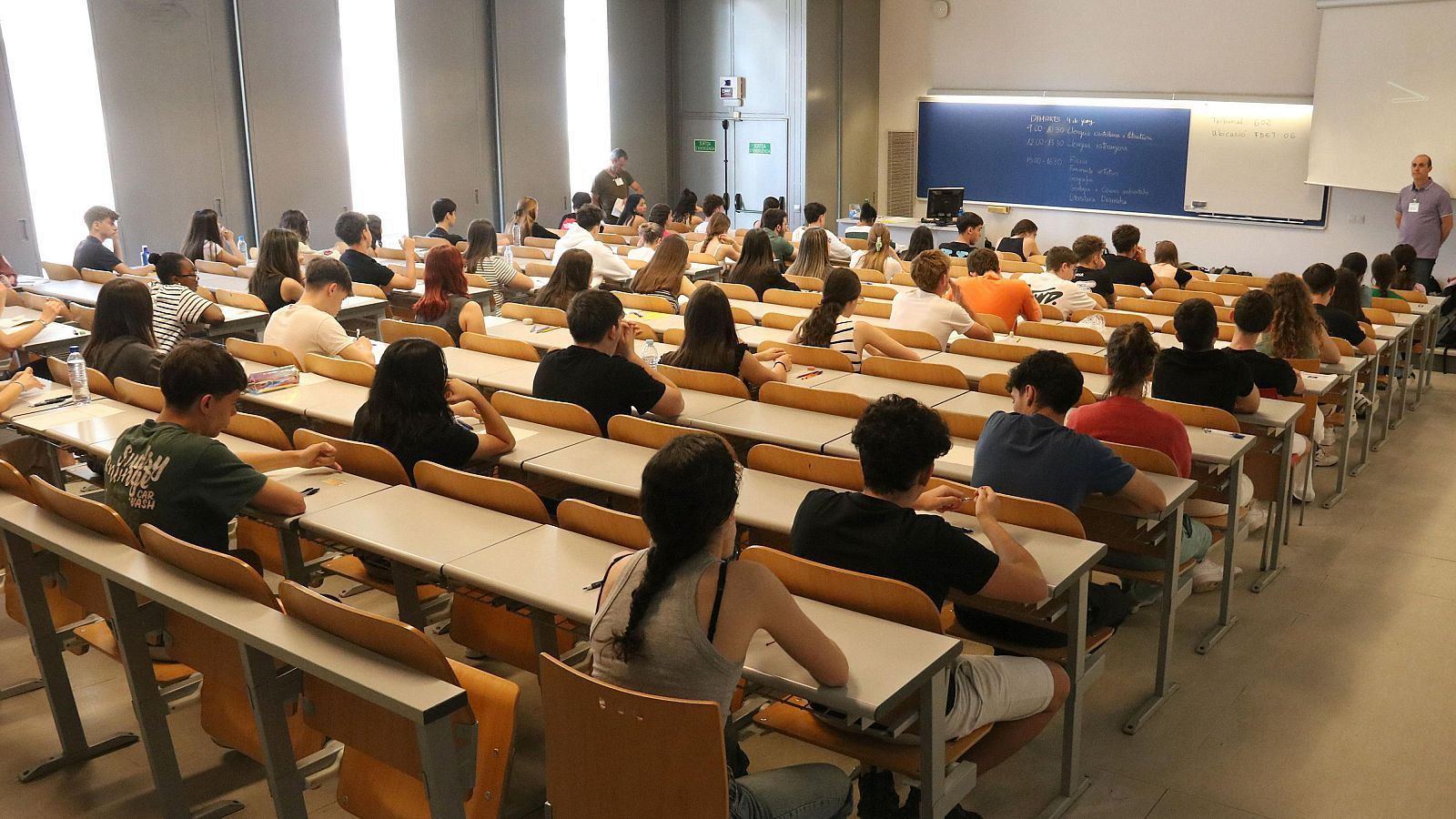 Jóvenes concentrados en un examen en un aula luminosa. Un supervisor supervisa desde el frente, junto a una pizarra y pantalla.