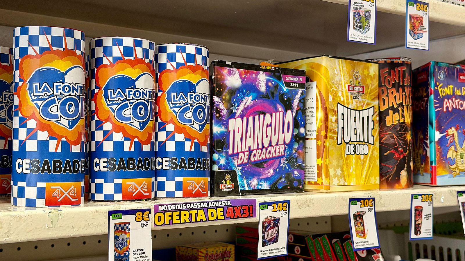 Estant de botiga amb focs artificials: cilindres "LA FONT COI", caixes "TRIANGULO DE CRACKER", "FUENTE DE ORO", "FONT BRUIXA del N...", "FONT del ANTONI" i cartell "NO DEIXIS PASSAR AQUESTA OFERTA DE 4X3!".