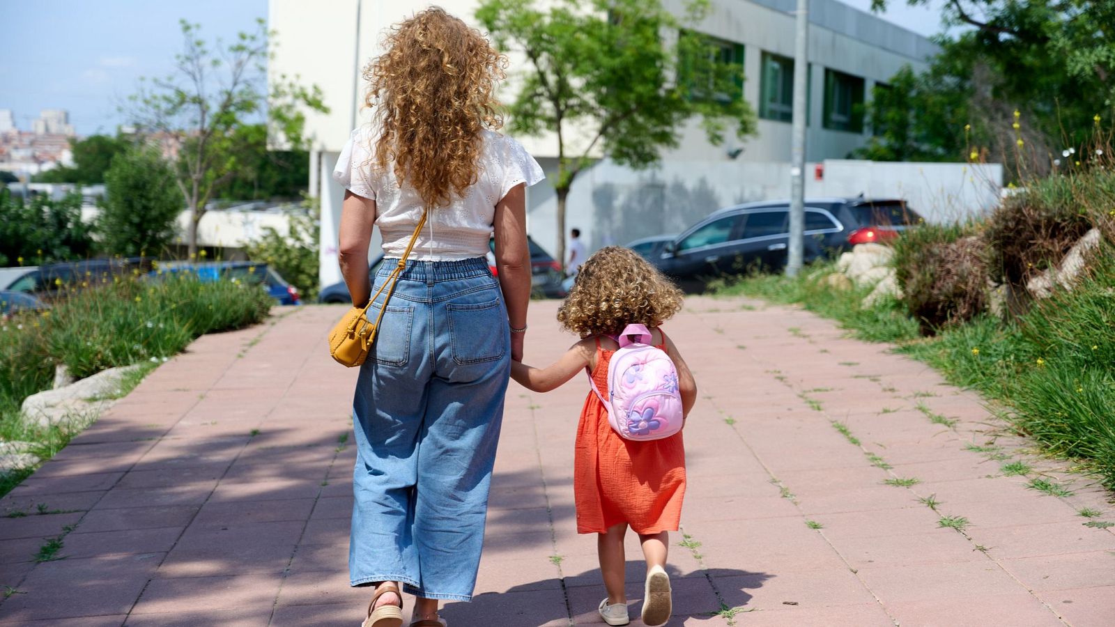 Mujer y niña pasean de la mano por un sendero soleado. Ella lleva camiseta blanca y vaqueros, la niña vestido naranja y zapatillas. Fondo urbano con edificios y cielo azul.