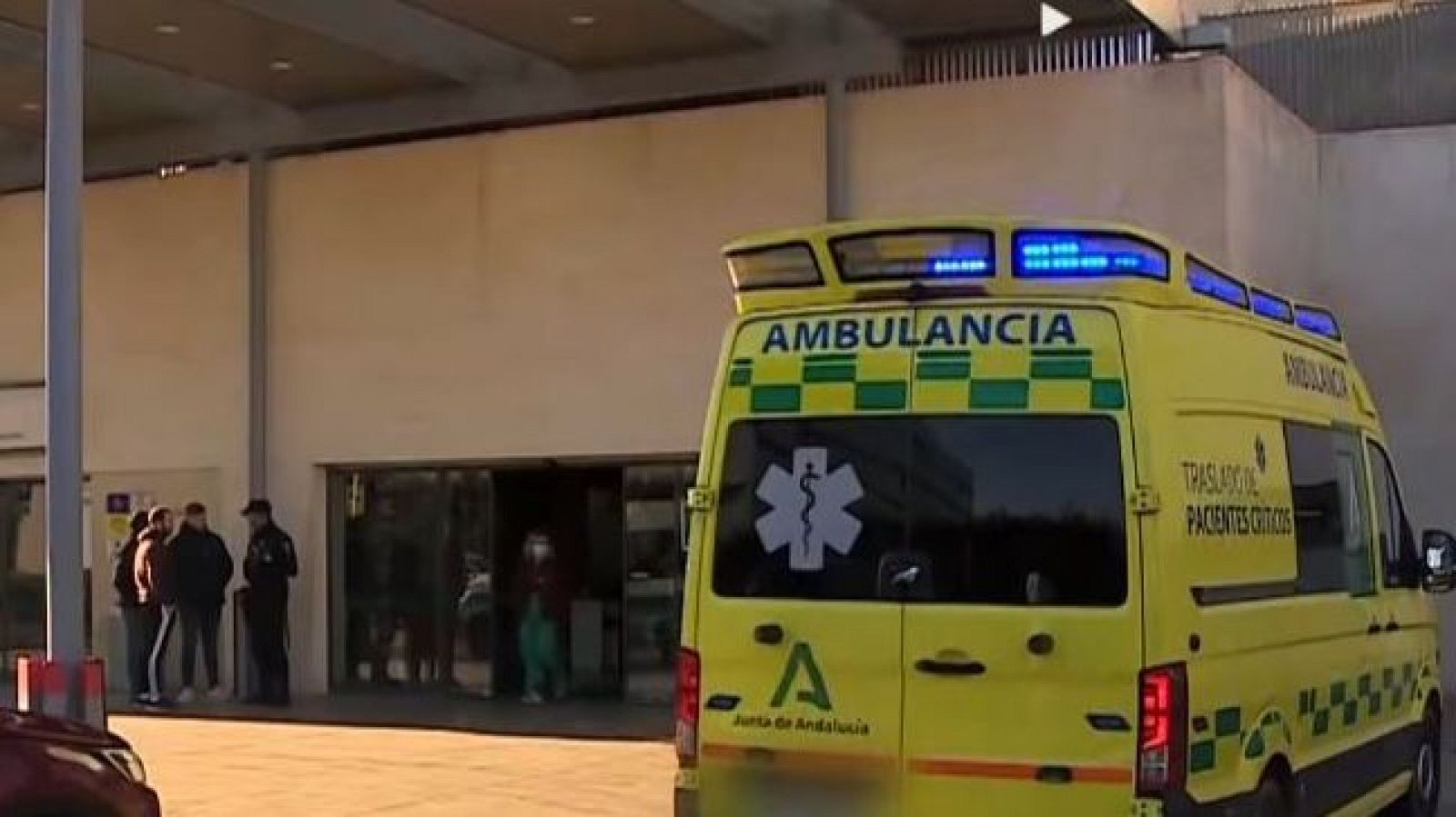 Ambulancia amarilla con franjas verdes y blancas, luces azules encendidas, frente a edificio médico. "AMBULANCIA", "TRASLADO DE PACIENTES CRÍTICOS", "Junta de Andalucía" visibles.