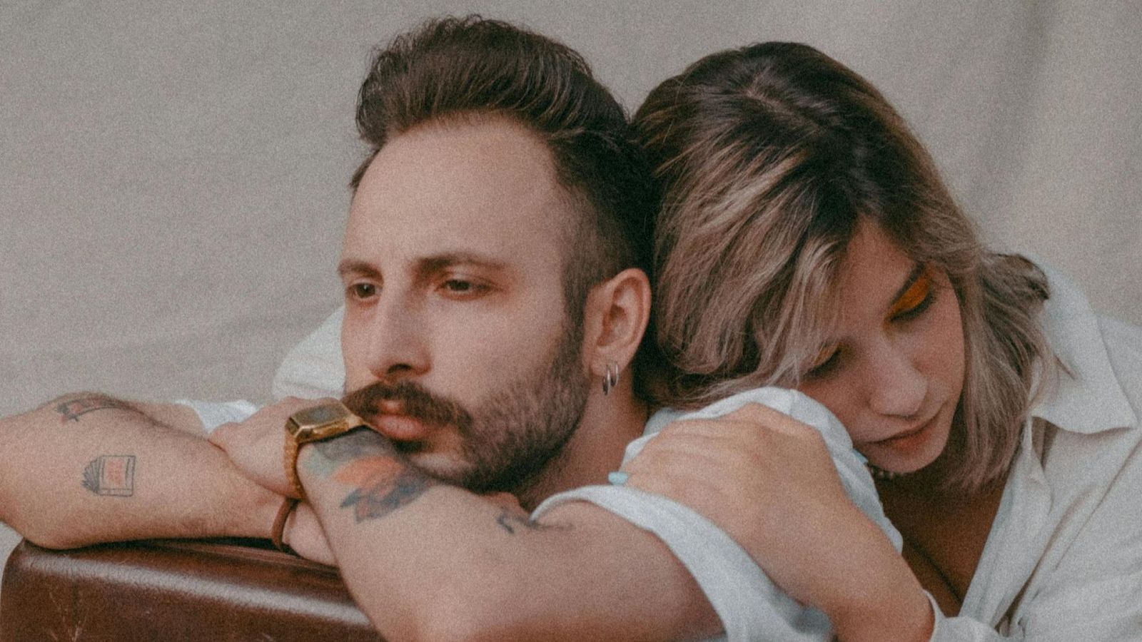 Primer plano de una pareja en pose íntima y reflexiva. Él, con barba y tatuajes, mira pensativo; ella, con maquillaje vibrante, apoya su cabeza en él. Fondo desenfocado.