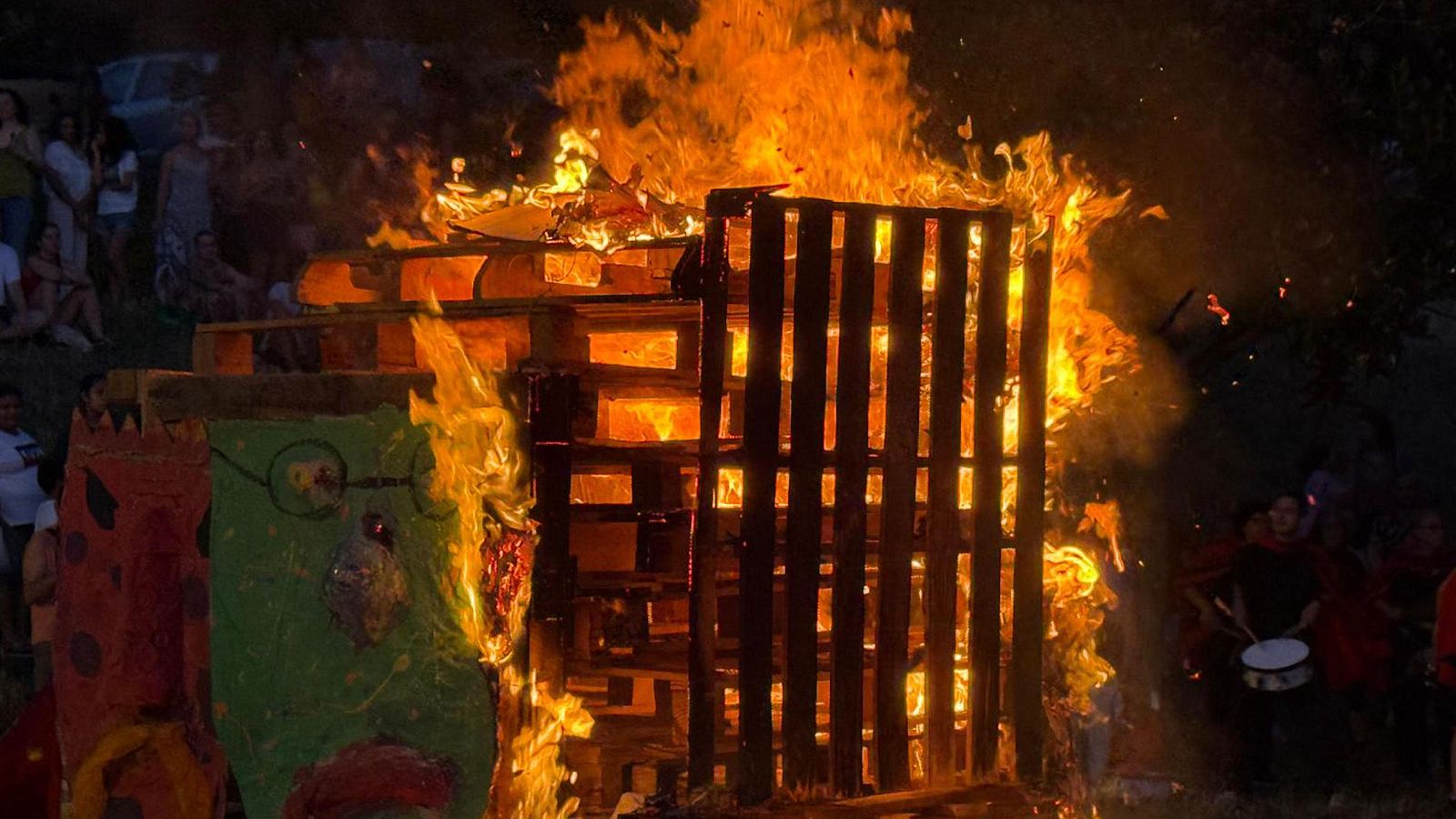 Hoguera nocturna de palés de madera con figuras verdes y rojas consumidas por las llamas, rodeada de espectadores y una persona con tambor.
