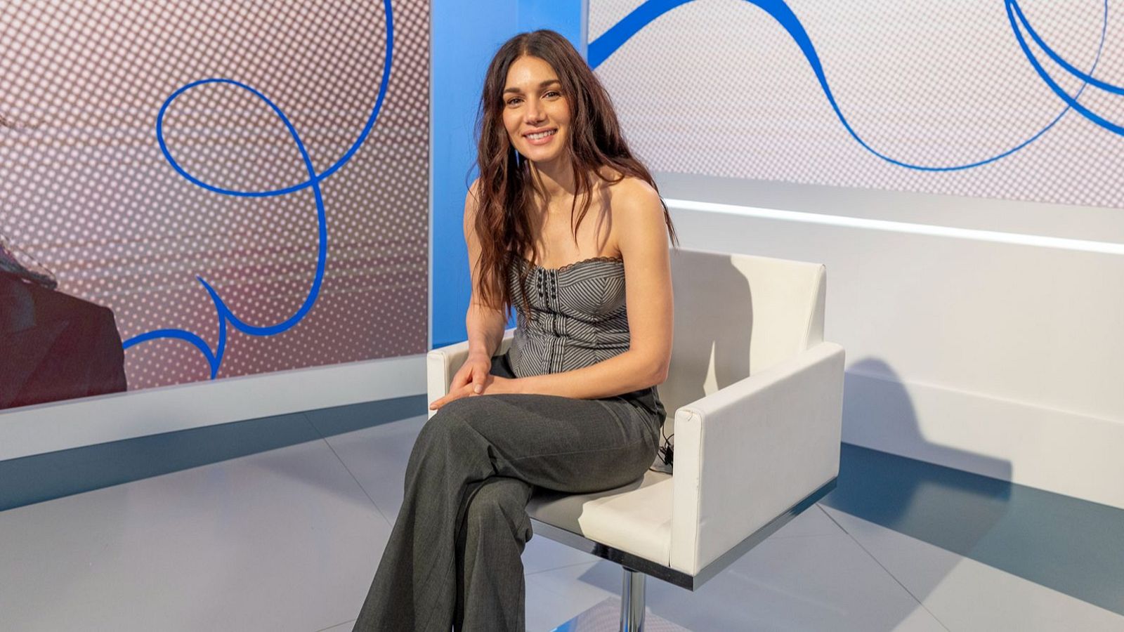 Mariela Garriga, con top de rayas y pantalones grises, sonríe sentada en un sillón blanco en un estudio con paneles decorados.
