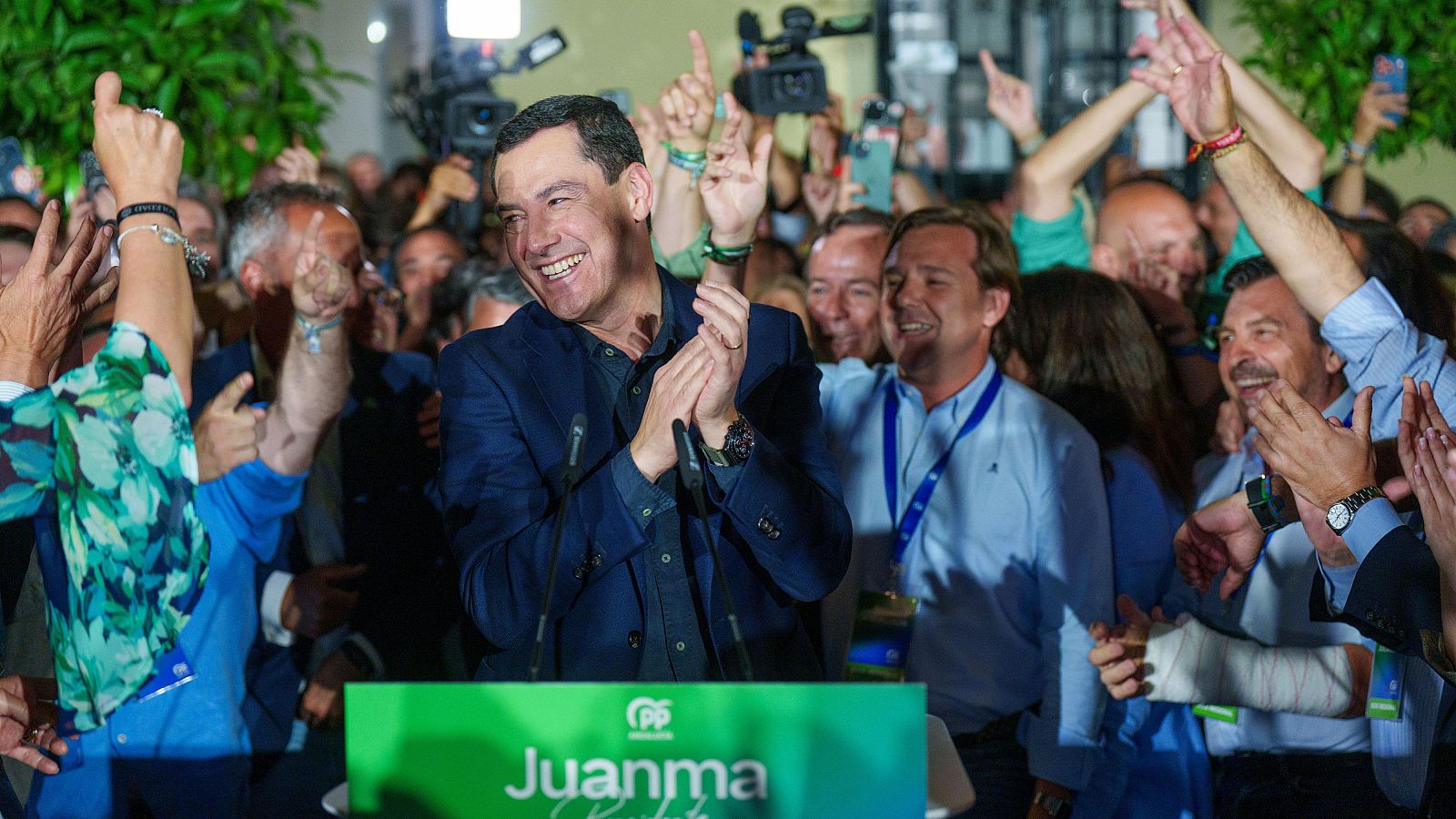 Juanma Moreno Bonilla, presidente de la Junta de Andalucía, sonríe y aplaude en un evento público. Lleva chaqueta azul marino y camisa a juego. Podio verde con logo PP y "Juanma" en blanco.