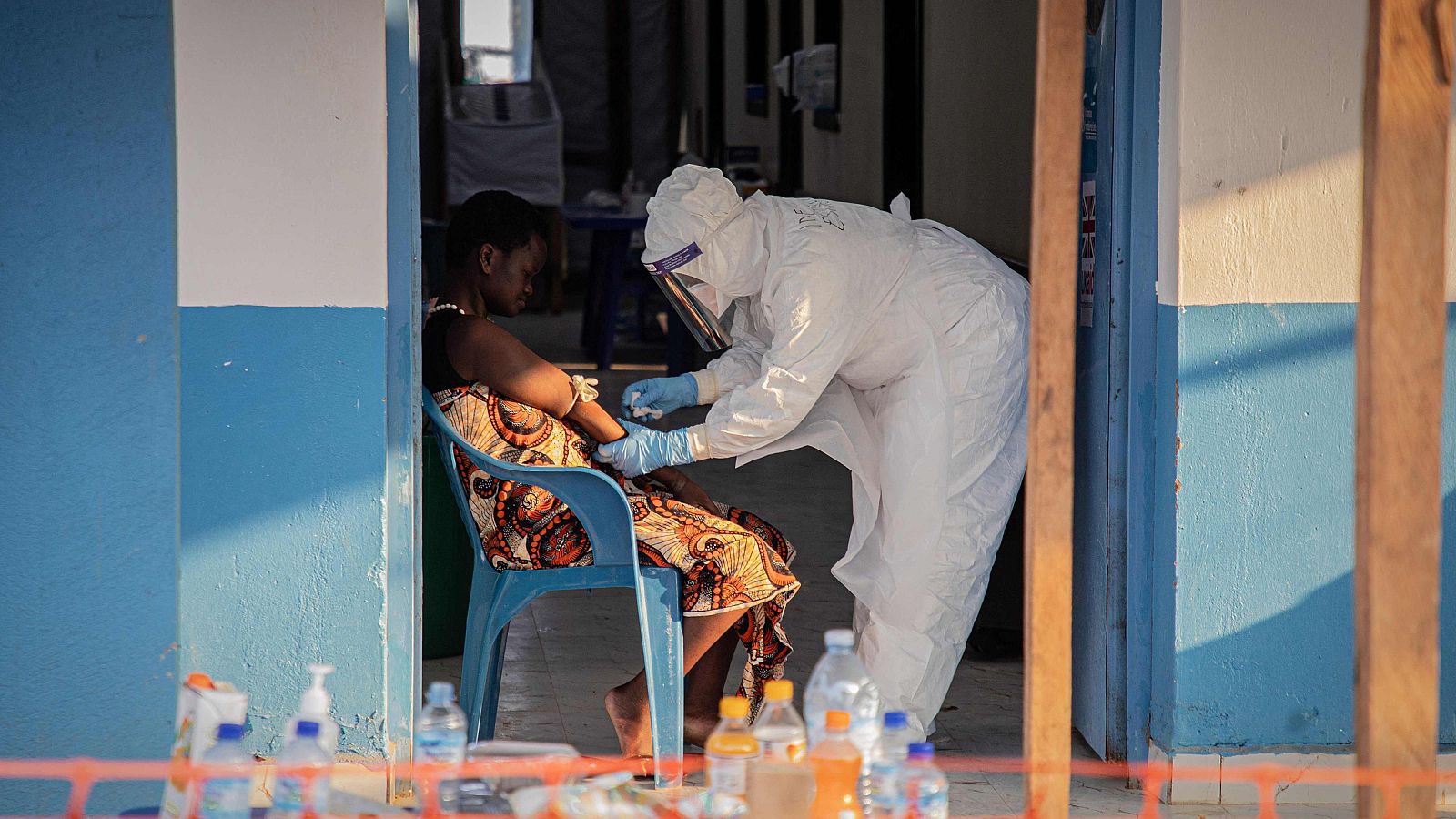 Trabajador sanitario con EPP atiende a paciente en silla azul, realizando procedimiento en su brazo. Botellas y dispensador de gel en primer plano. Bunia, RDC, 23 de junio de 2026.