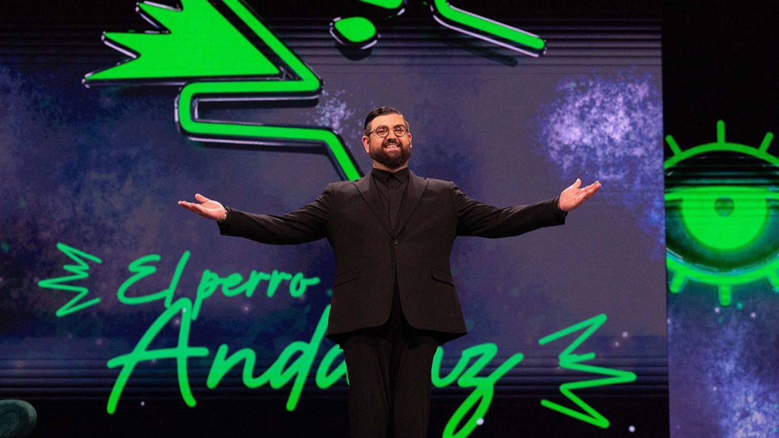Manu Sánchez, con traje oscuro y gafas, sonríe en un escenario con fondo digital azul-morado y el texto "El perro Andaluz" en verde neón.