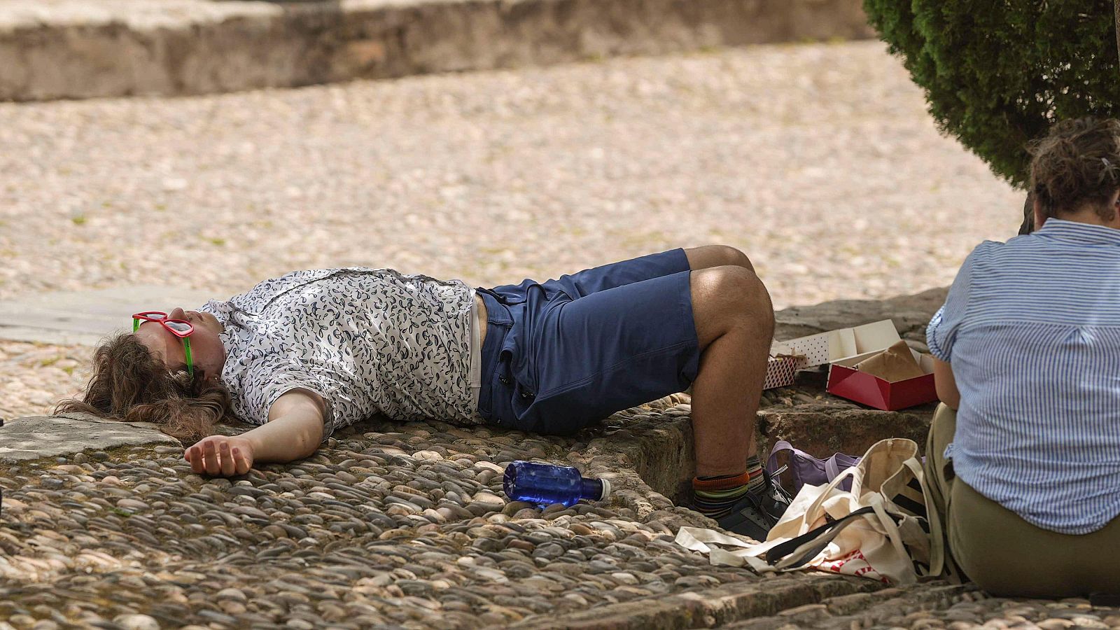 Dos personas buscan alivio del calor en un día soleado, una tumbada en el suelo y la otra sentada a la sombra.