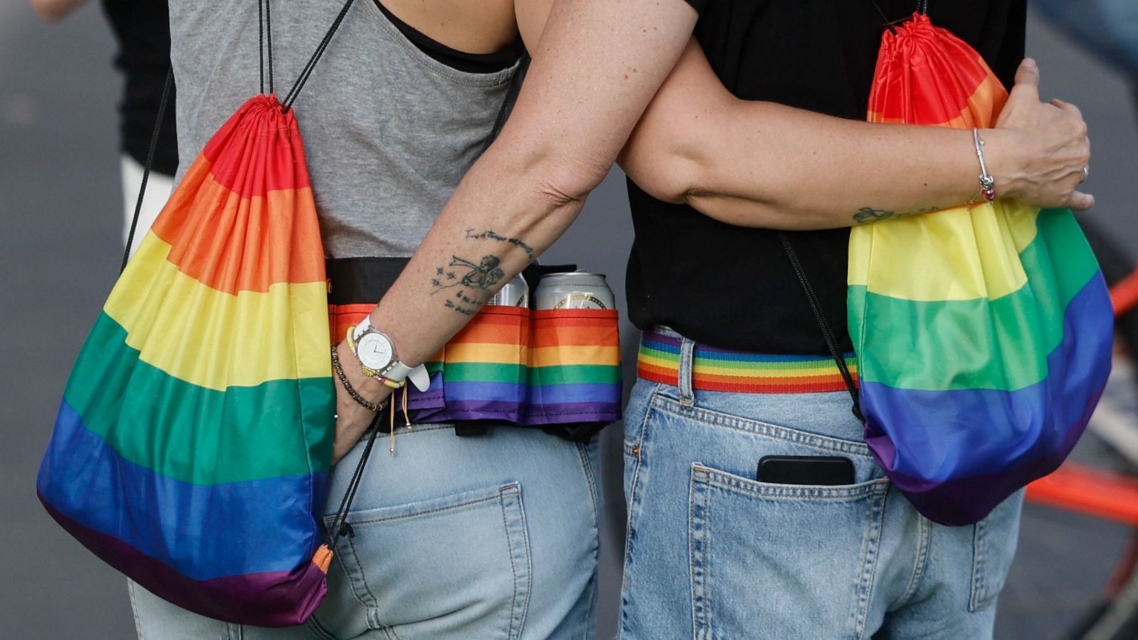 Manifestación con dos personas de espaldas, luciendo mochilas arcoíris y jeans. La persona de la izquierda muestra un tatuaje y riñonera con latas; la de la derecha abraza a su acompañante.